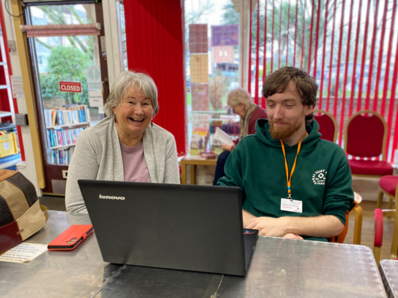 A young male volunteer sitting at a table with a laptop in front of them. They are both looking towards the camera and smiling.