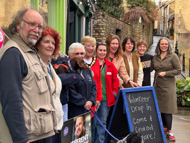 A group of people standing outside on a high street, they are smiling at the camera, one is holding a dog and there is an a-frame in front of them saying 'volunteer drop in'