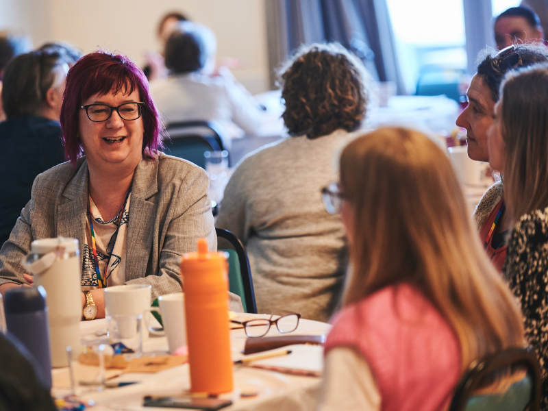 Photograph of two women sat at a table at an event.. One is face on and smiling, the other has her back to the camera. There are more people in the background. There are papers, drinking glasses and a bright orange bottle on the table.