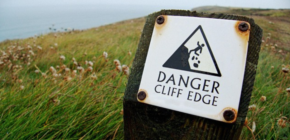 Photograph of a sign with the words 'Danger cliff edge' on a grassy cliff edge