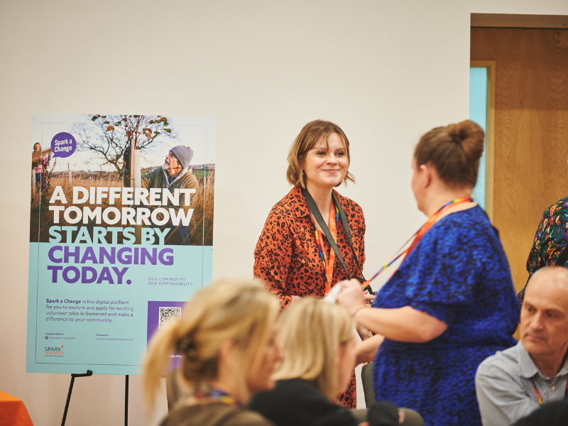 Photograph of two women in conversation at an event, there is a Spark a Change post to their right and two people seated in the foregorund.
