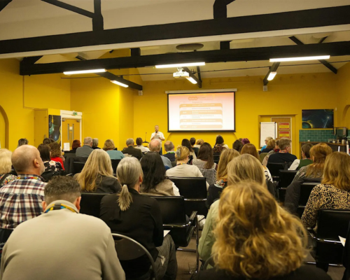 A photo from a previous, in-person research meeting; a room of people are sat and listening attentively to the speaker