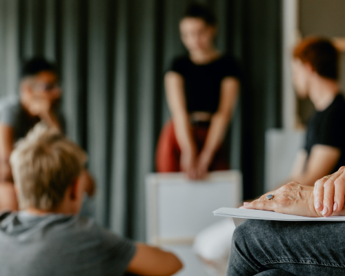 An image of a group of young people stood and sat in a circle, in conversation. They are out of focus. In the foreground are a pair of hands belonging to an older person, resting on a clipboard on their lap. The person is cropped so only this is visible.