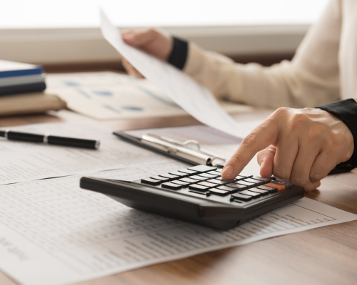 A close up photo of a person sat at their desk. They are holding a sheet of paper and using a calculator.