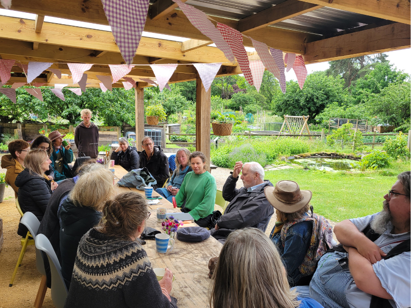 Photograph of a group of people sitting around a large table. They are in an outdoor setting with bunting overhead.