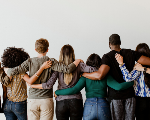 A photo depicting a diverse group of people, photographed from behind. They are stood in a line and have their arms around each other.