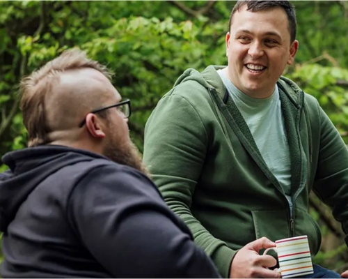 An image of two men, sat outdoors, drinking tea from mugs and smiling