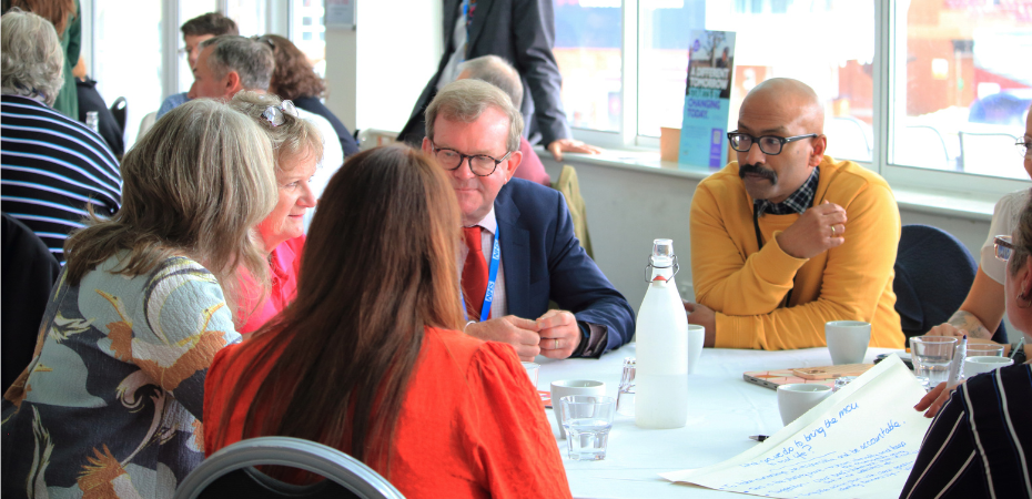 Photograph of a group of people sat at a table having a discussion in a conference setting