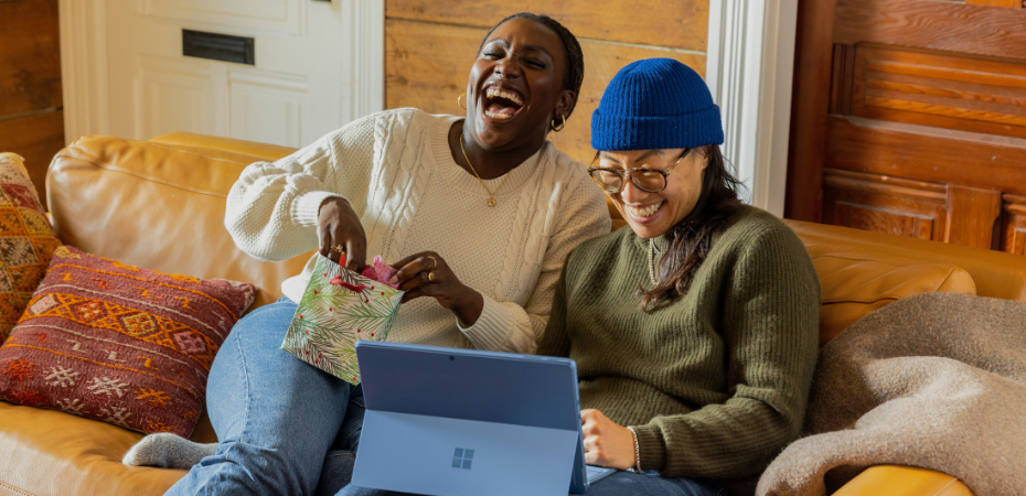 Two women sat on a sofa together. One is using a laptip and smiling, the other is holding a small gift bag and throwing her had back in laughter.