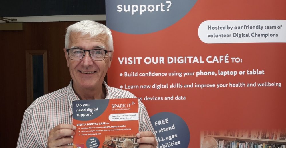 A photo of a man posing next to a Spark iT pull-up banner, and holding a Spark iT flyer. He is looking at the camera and smiling.