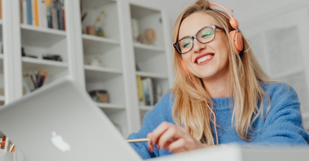 Photograph of a young woman using a laptop, she is smiling and wearing bright orange earphones.