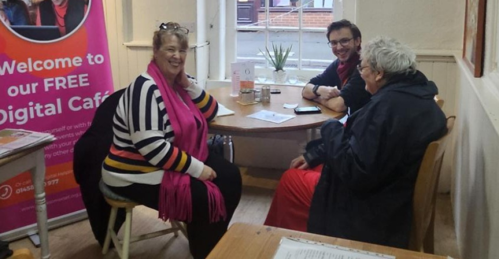 Lynne, featured in the article sitting at a table with a young male volunteer and older female client. They are smiling at the camera. There is a Spark iT promotional banner behind Lynne.