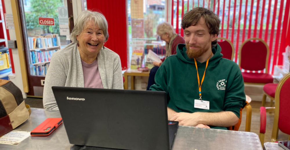 Sue a visitor to the Digital Cafe featured in the story sitting alongside volunteer Digital Champion Morgan. They have a laptop in front of them on the table and Sue is smiling warmly at the camera.