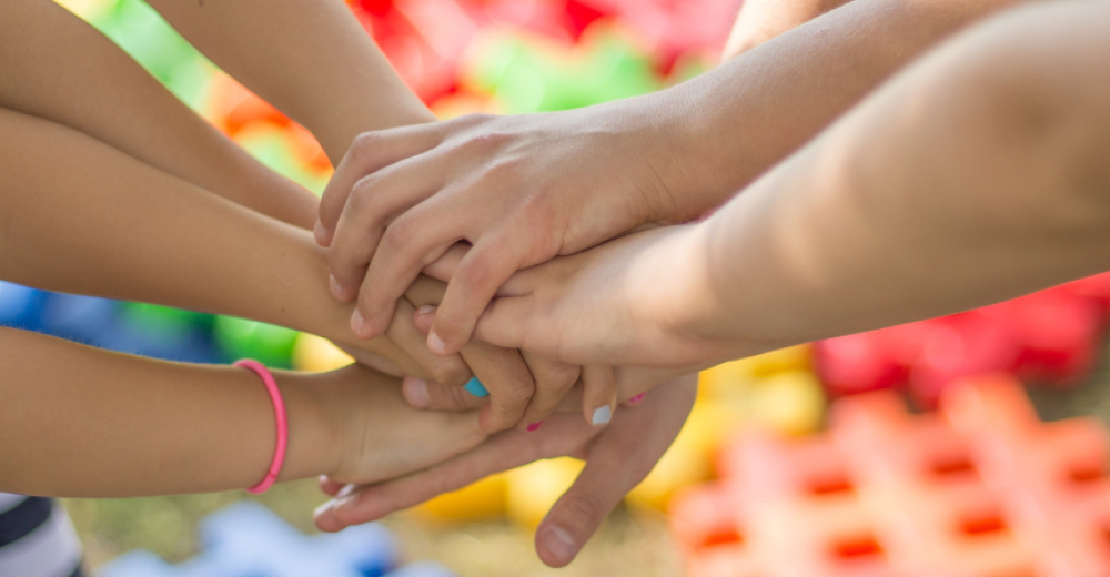Childrens holding hands in a circle with only their hands and forearms in shot, some have painted fingernails and there are colourful lego type blocks in the background.