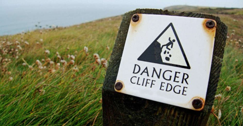Photograph of a sign with the words 'Danger cliff edge' on a grassy cliff edge