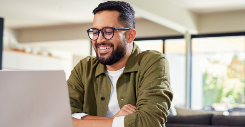 Photogrpaph of a man sitting at a table using a laptop, he wearing black framed glasses and a green shirt. His arms are folded and he is smiling.