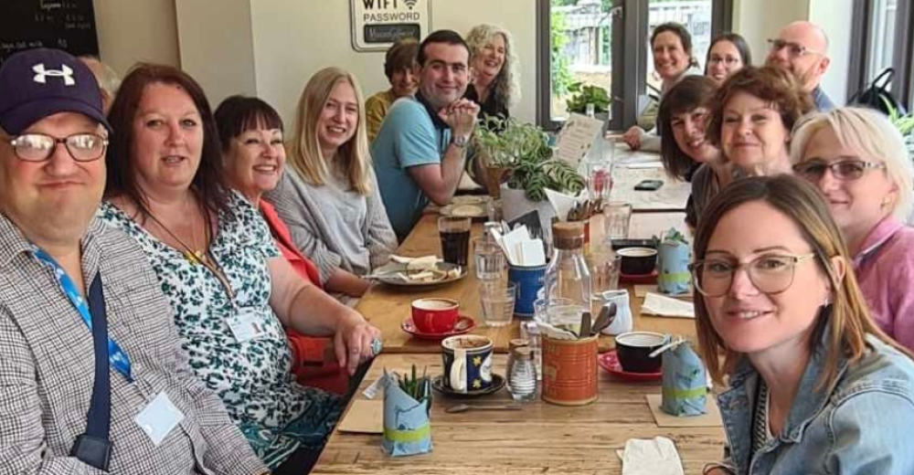 A photograph of a groups of volunteers sitting at a table in a cafe. They are looking towards the camera and smiling. Anna is at the front on the right.