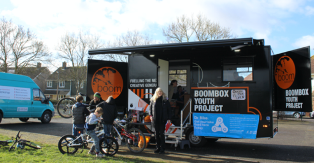 Photograph of a woman and children on bikes are standing in front of the Boombox youth project on wheels.