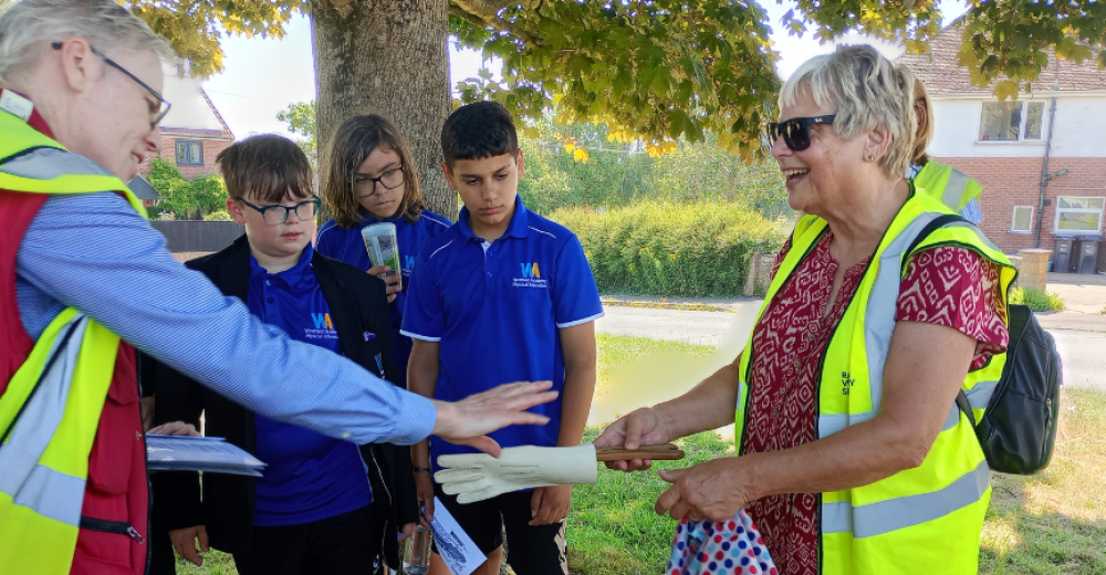 A photograph of a man, wearing a high visibility vest handing an artefact to a woman in a high visibility vest. They are in a part and overlooked by three young school boys.