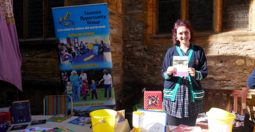 A member of the Taunton Opportunity Group standing at a table with donation buckets and leaflets. She is outside in the sunshine and smiling.