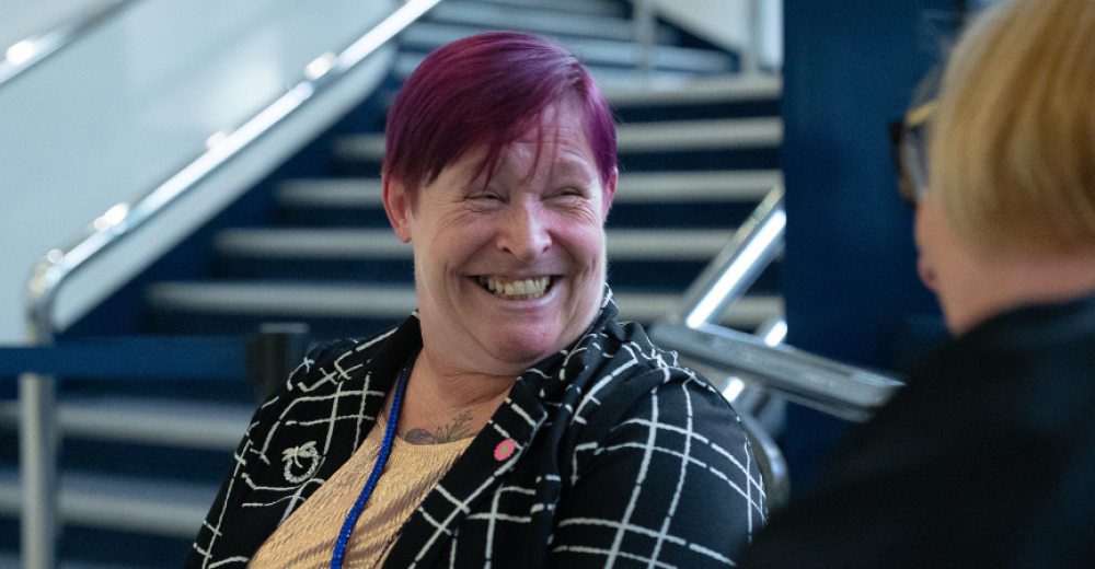 A close up photograph of two women in conversation. One is face on and smiling, the other has her back to the camera.