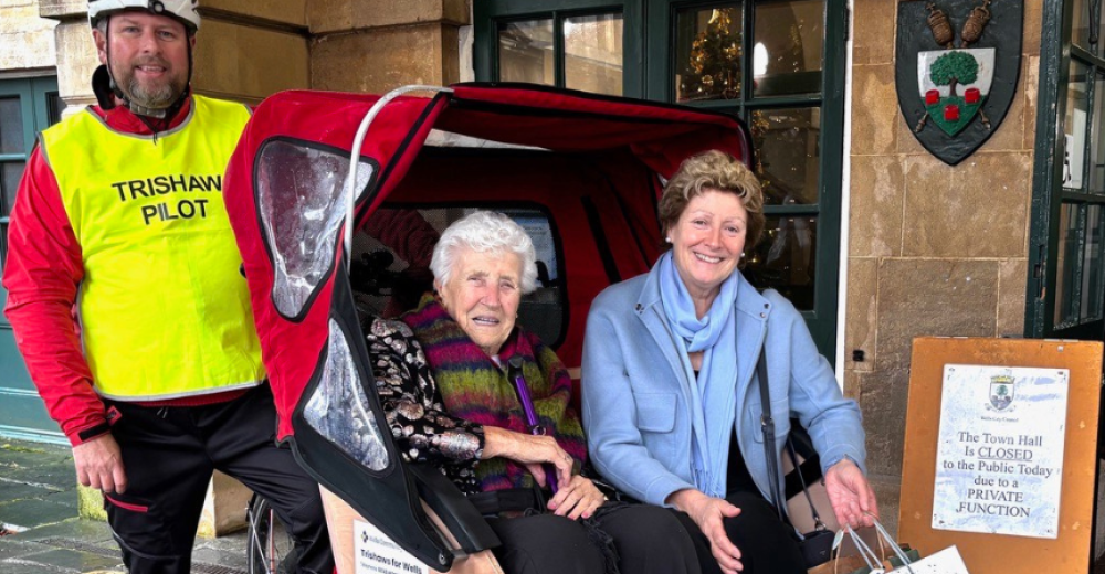 A picture of two ladies sat in the seat of a trishaw with the pilot stood behind them. They're all smiling at the camera.